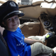 Boy in blue shirt and khaki shorts with a black hat smiling at the camera