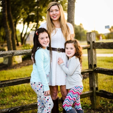 Mother in white dress, smiling with her two young daughters alongside a wooden fence