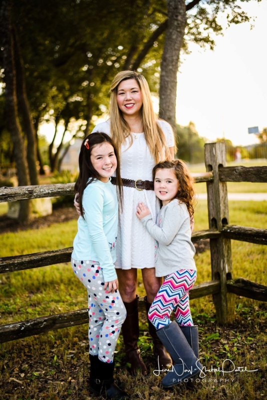 Mother in white dress, smiling with her two young daughters alongside a wooden fence