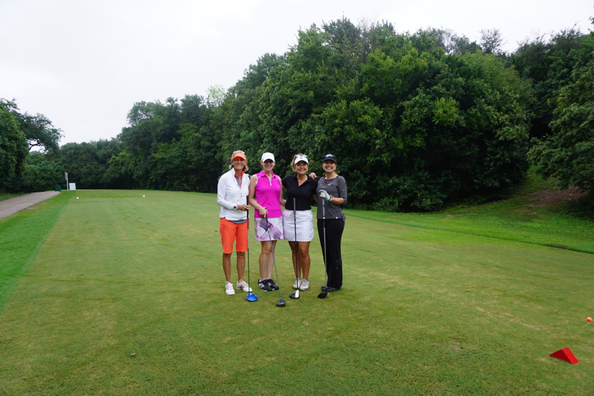 Four women standing together in a golf course holding golf clubs and smiling