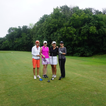 Four women standing together in a golf course holding golf clubs and smiling