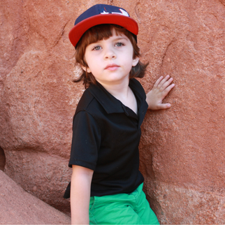 Boy in red and blue cap, black shirt, and green shorts against a rock