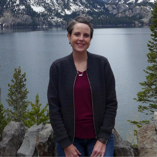 Woman in jeans, red shirt, and black hoodie with a lake behind her