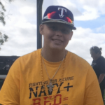 Boy in yellow shirt and texas rangers hat smiling at camera outdoors